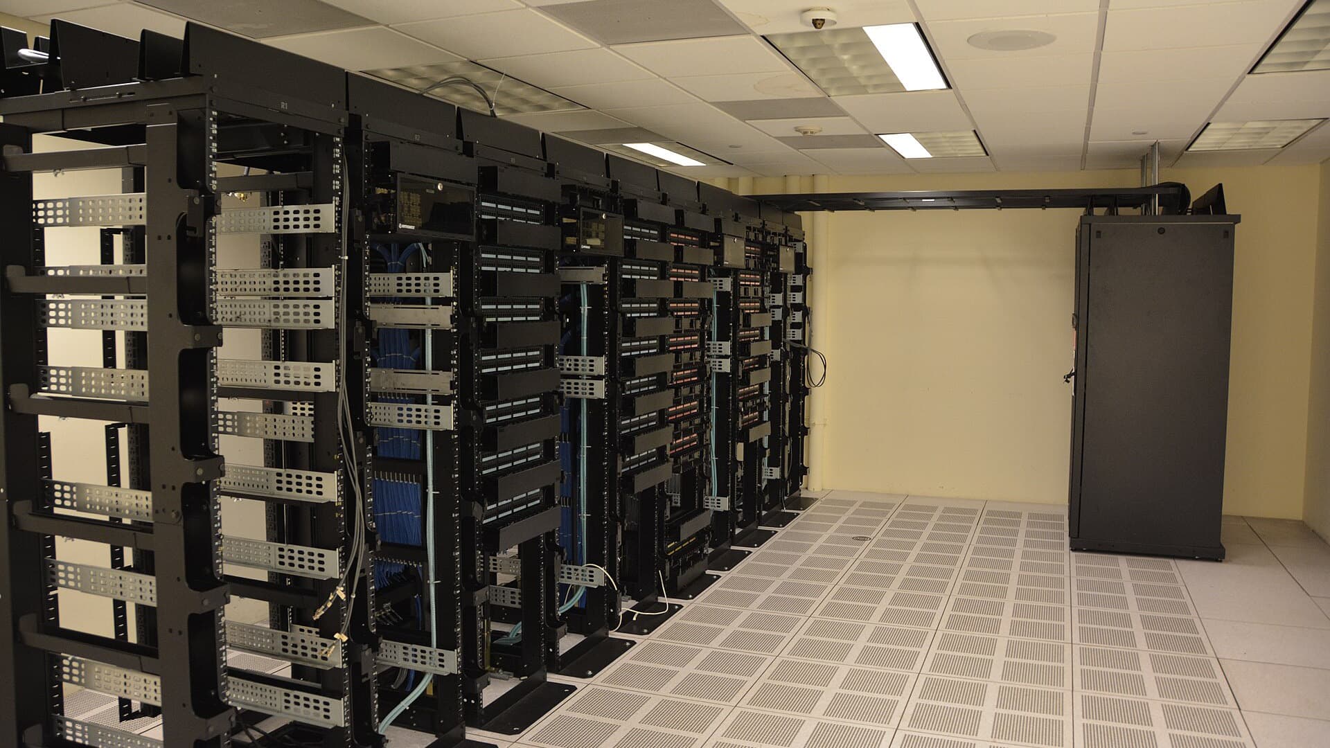 Rows of black server racks with network cabling in a brightly lit data center with a raised white floor.