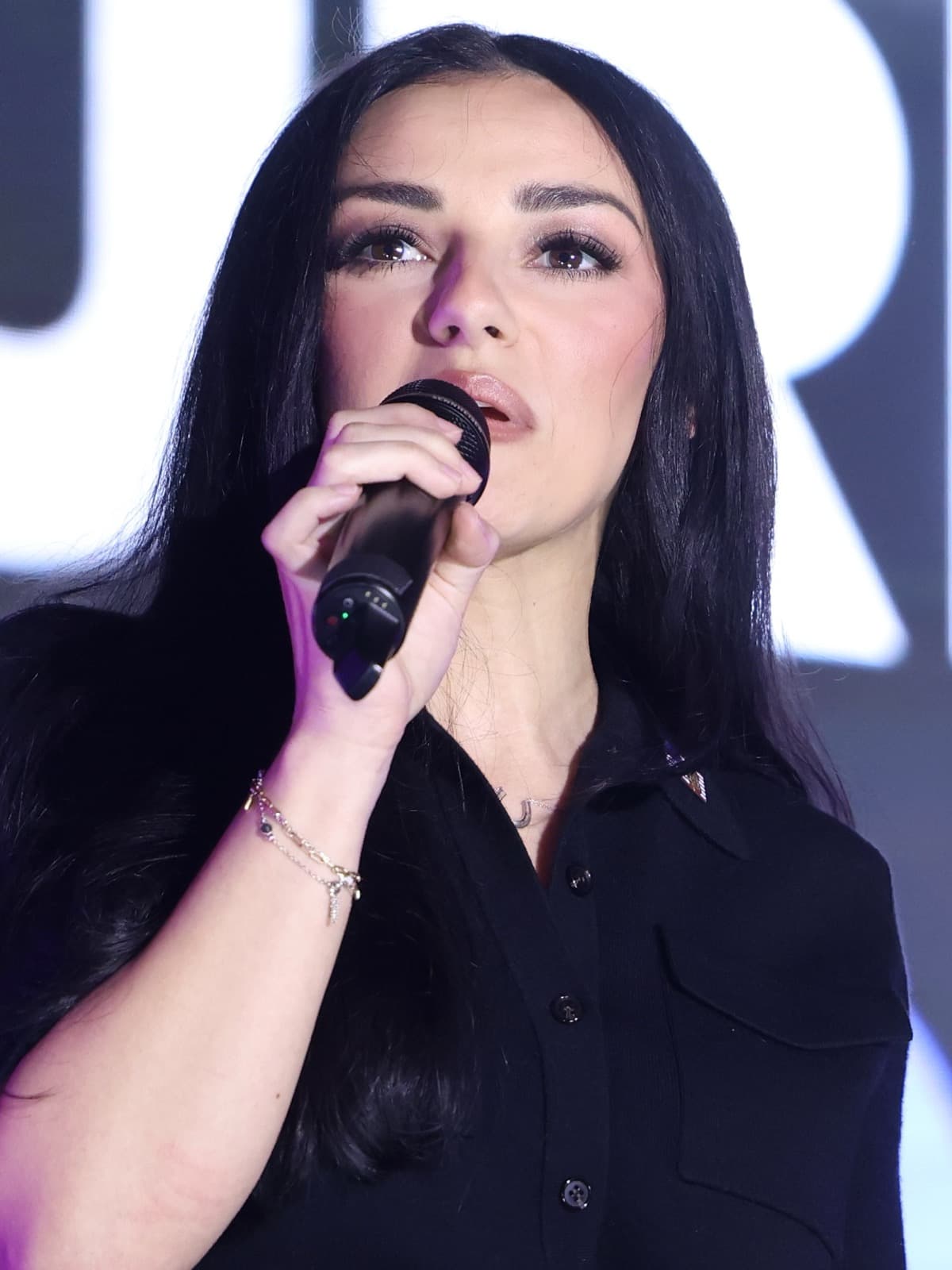 Alexis Wilkins, a woman with long dark hair in a black collared top, singing into a handheld microphone on stage.