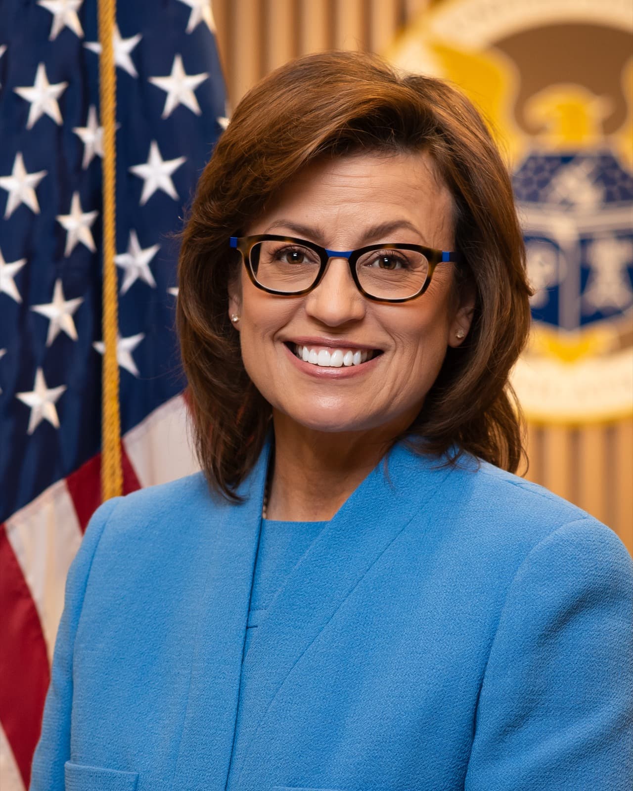 Official portrait of Anna M. Gomez, a smiling woman with shoulder-length brown hair and dark-framed glasses in a blue jacket, in front of a U.S. flag.