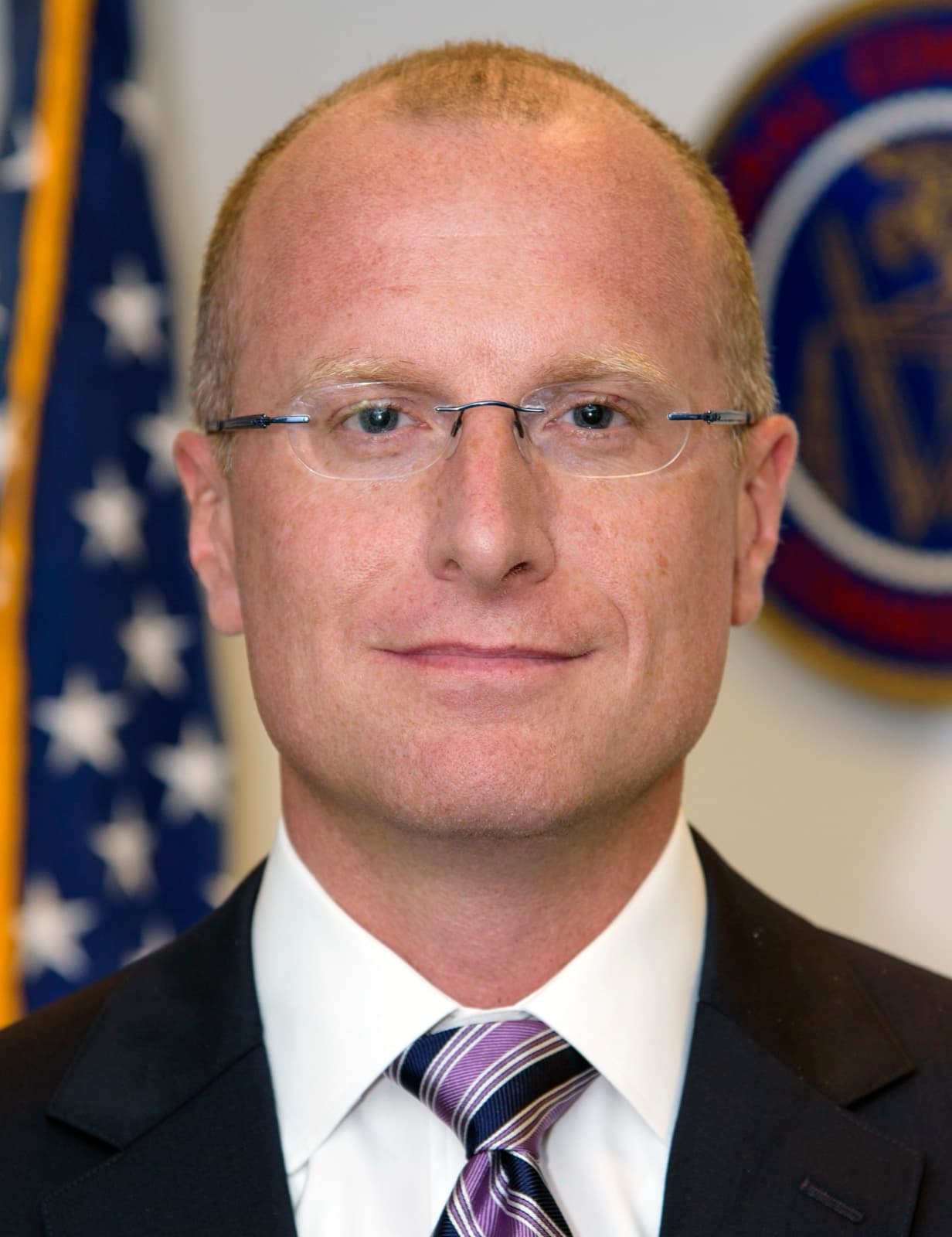 Official portrait of Brendan Carr, a man with short hair and rimless glasses in a dark suit, white shirt and purple striped tie, in front of a U.S. flag and the seal of the Federal Communications Commission.
