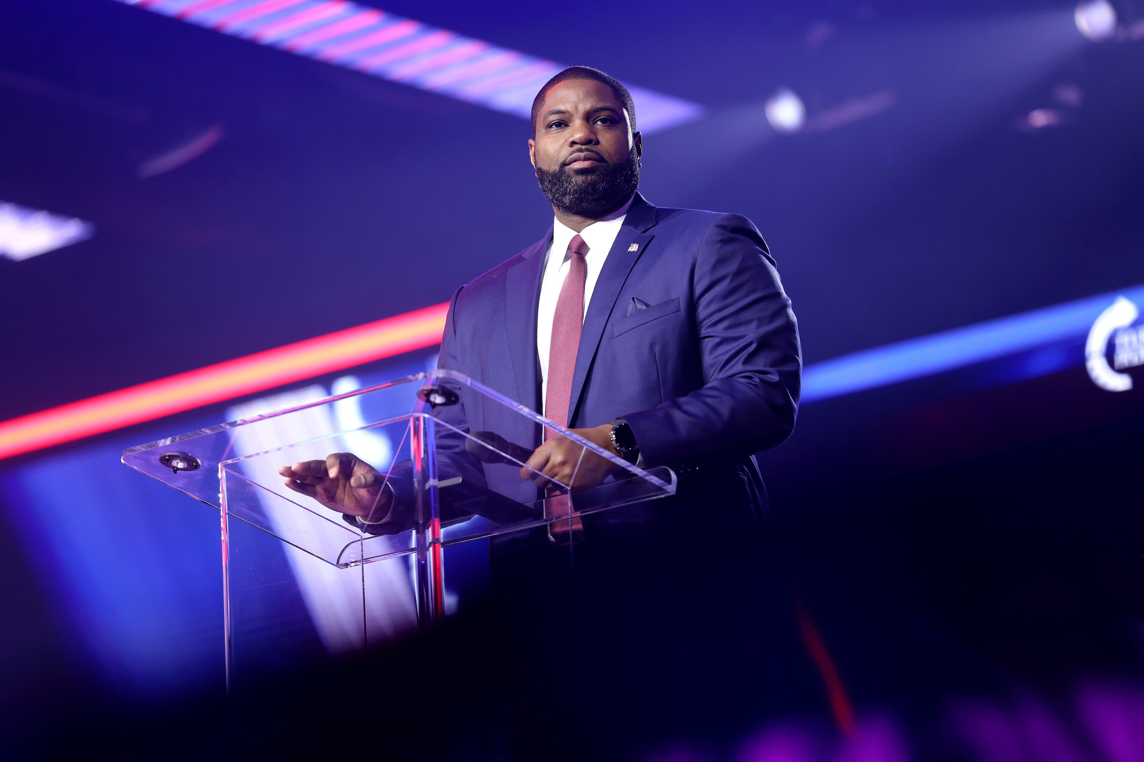Byron Donalds speaking at a lectern under red and blue stage lighting.