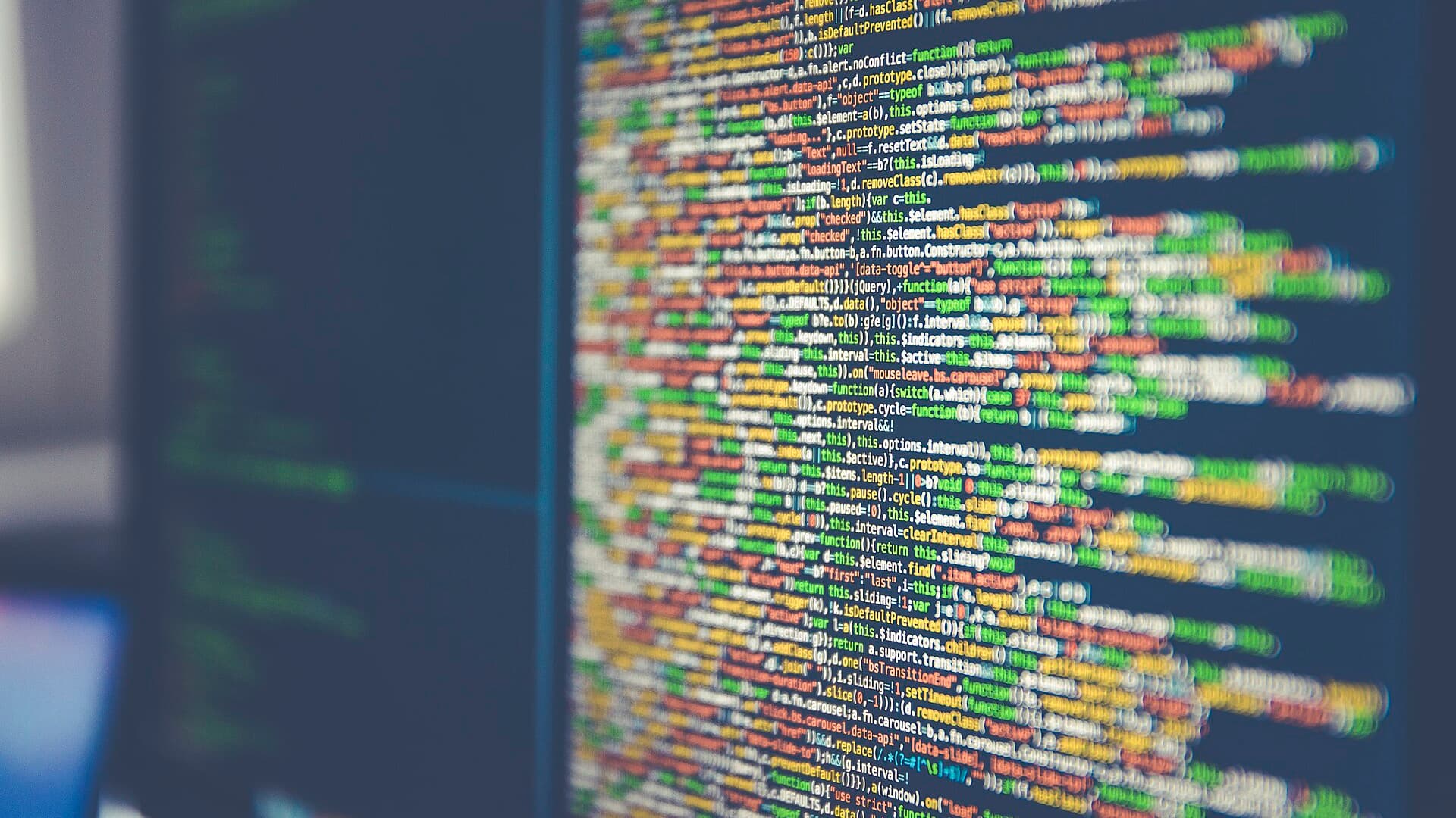 Close-up of lines of colorful program code on a computer monitor, with a second screen blurred in the background.