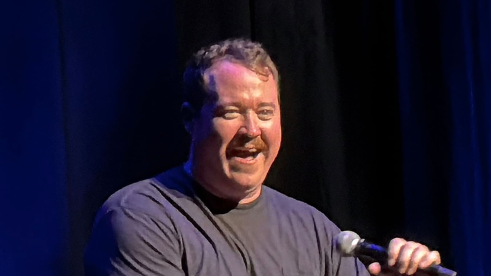 Shane Gillis holds a microphone while performing onstage.