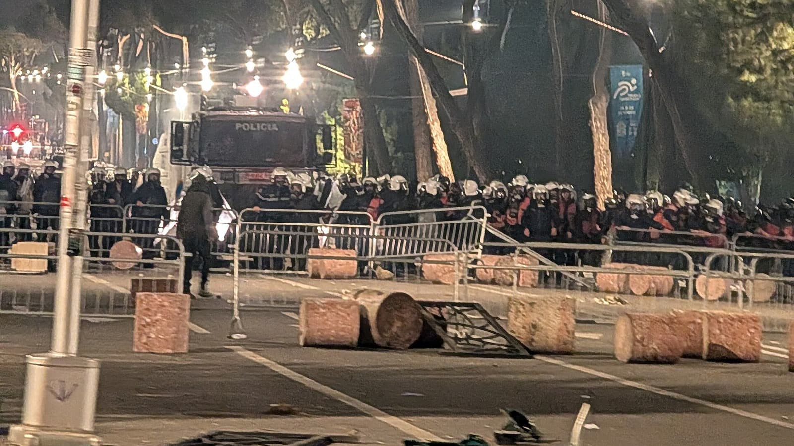 A line of police behind barriers on a street during protests in Tirana at night.