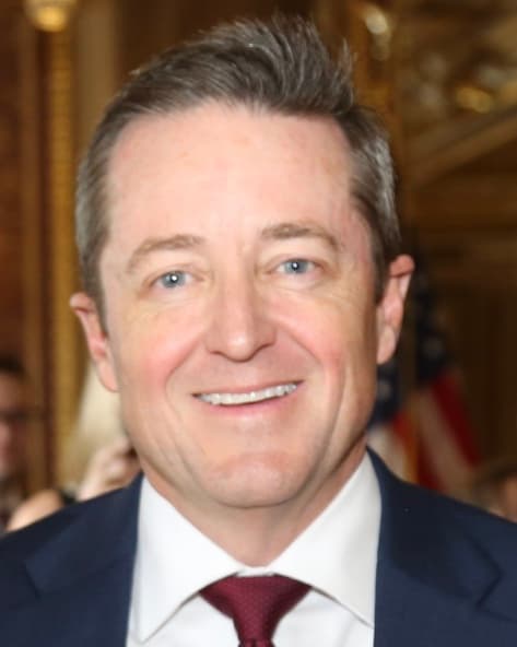 Derek Brown, wearing a dark suit and red tie, smiles in an ornate room with an American flag behind him.