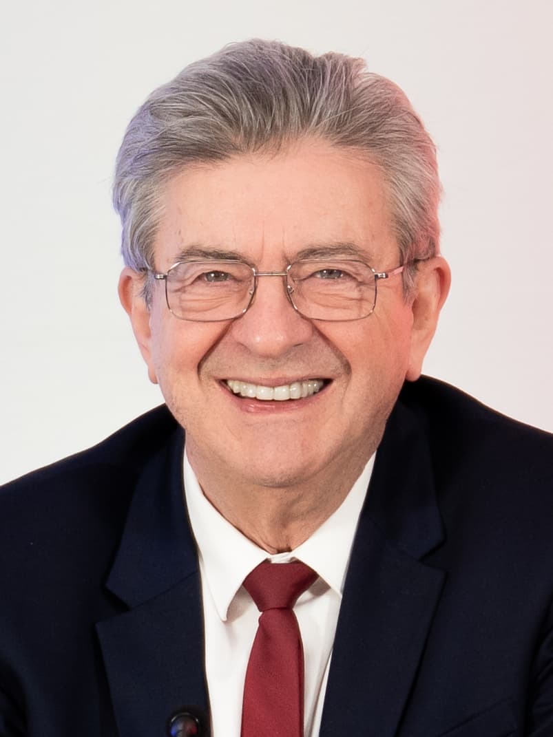 Jean-Luc Mélenchon smiling, in a dark suit and red tie, against a white background.