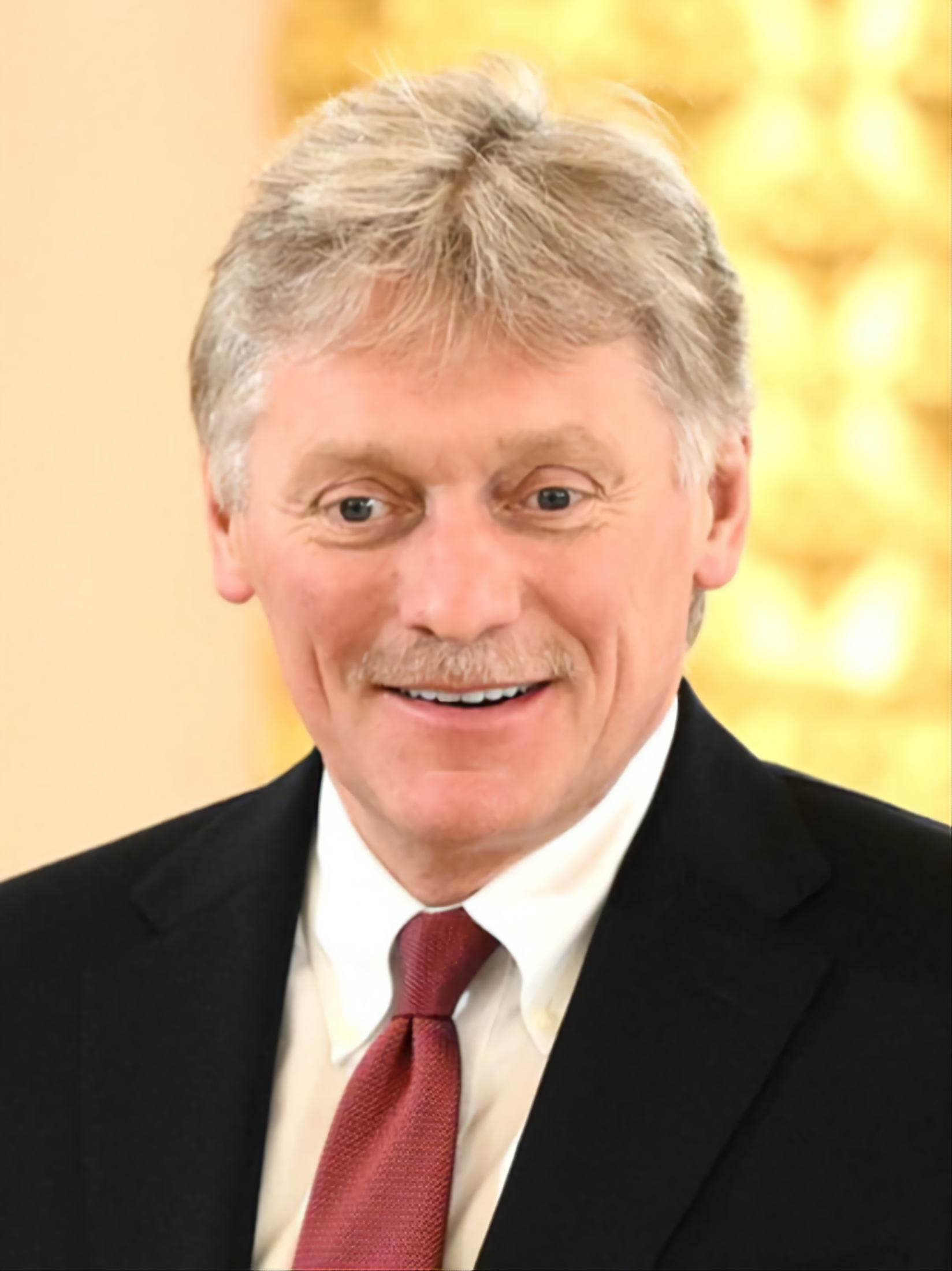 Portrait of Dmitry Peskov, a man with gray hair and a mustache, in a dark suit, white shirt and dark red tie.