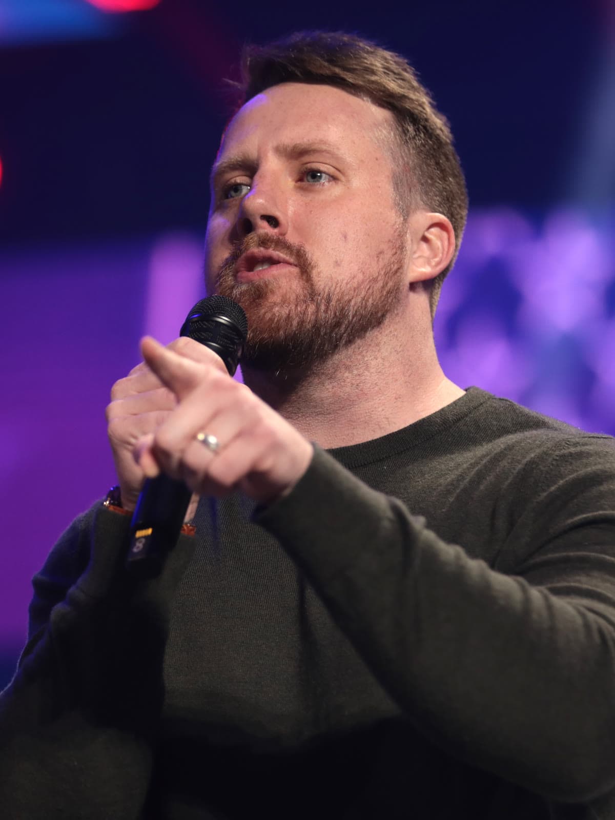 Elijah Schaffer, a bearded man in a dark green sweater, speaking into a handheld microphone on a stage.