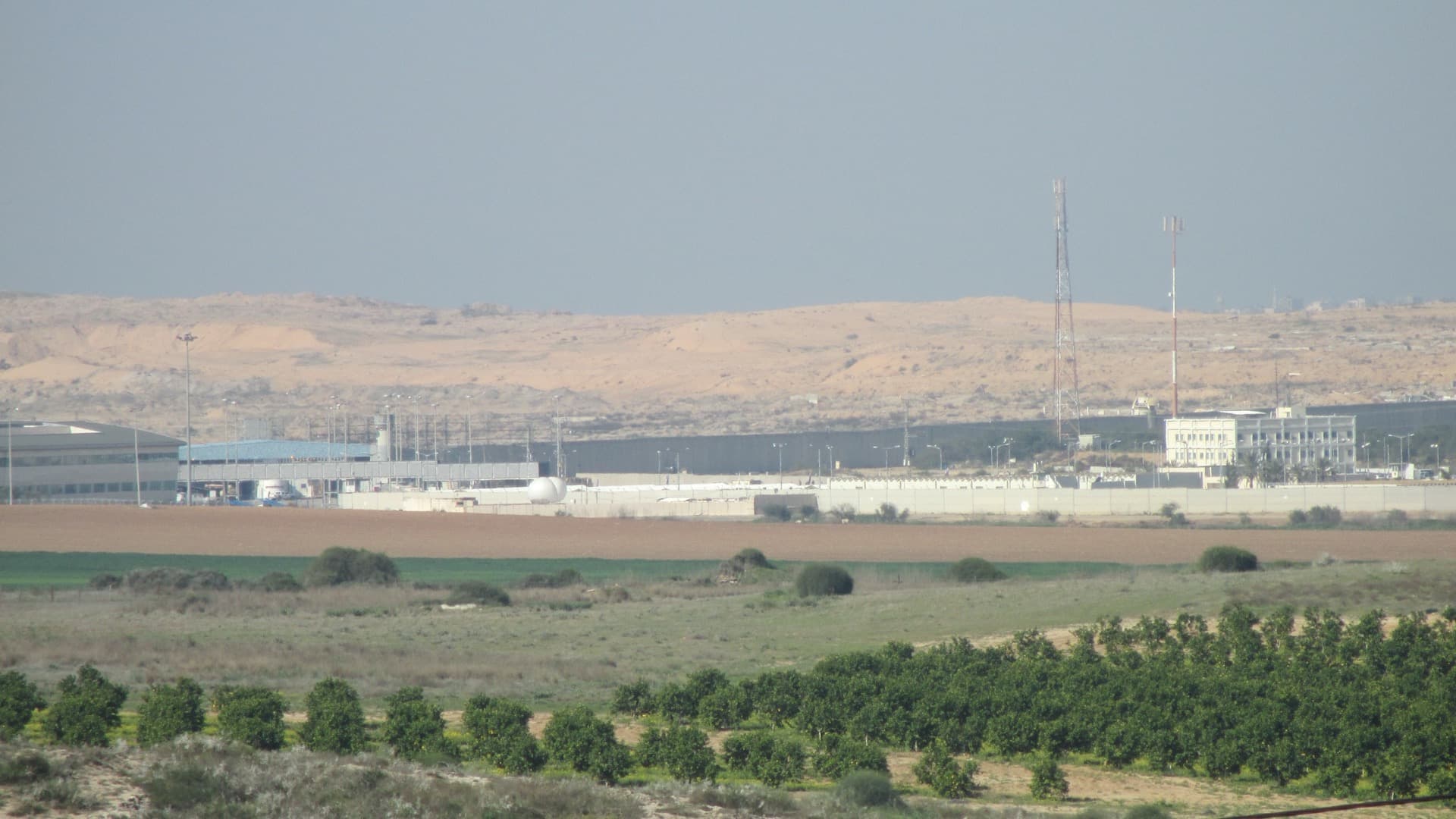 The Erez crossing between Israel and the northern Gaza Strip, seen across farmland and orchards from Kibbutz Erez, with sand dunes in Gaza behind it.