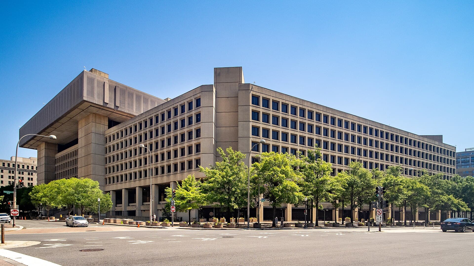 The J. Edgar Hoover Building, the FBI headquarters in Washington, a large concrete building with rows of windows, seen from a street corner under a clear sky.