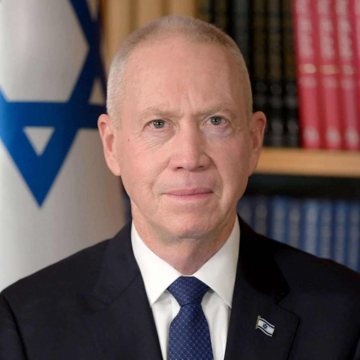 Yoav Gallant in a dark suit, white shirt and blue tie, seated in front of an Israeli flag and a bookshelf.
