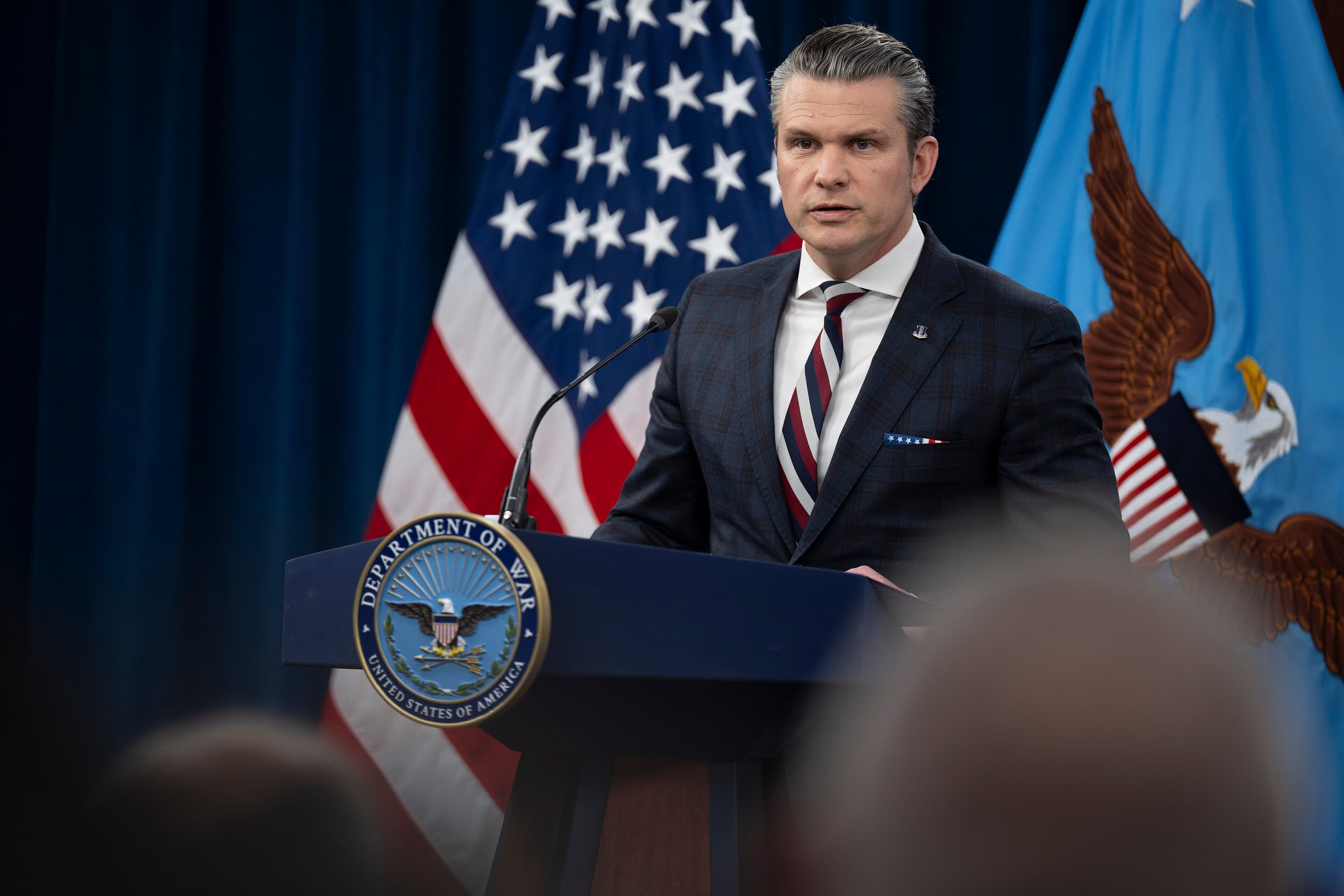Pete Hegseth speaking at a Pentagon briefing with flags behind him.