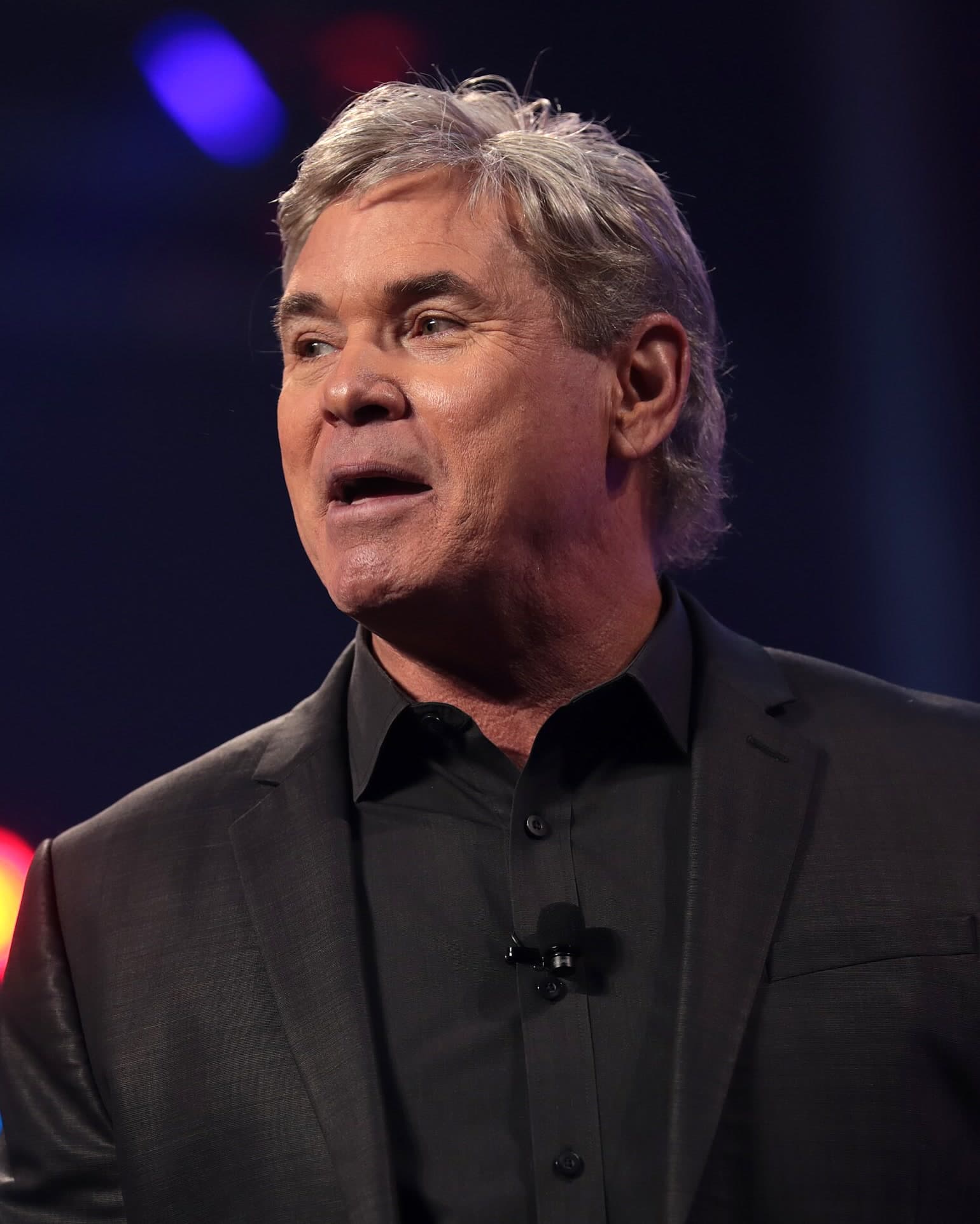 Jack Hibbs, a man with gray hair in a dark jacket and shirt, speaking into a lapel microphone on a stage.