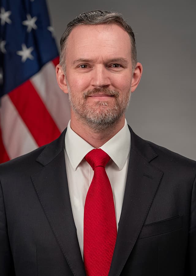 Official portrait of U.S. Trade Representative Jamieson Greer in a dark suit and red tie in front of an American flag.