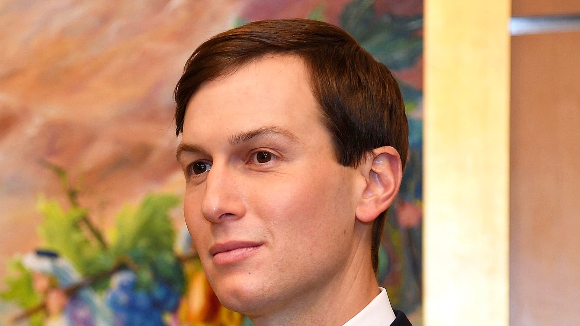 A close-up portrait of Jared Kushner in a dark suit and white shirt, looking to the left in front of a colorful painting.