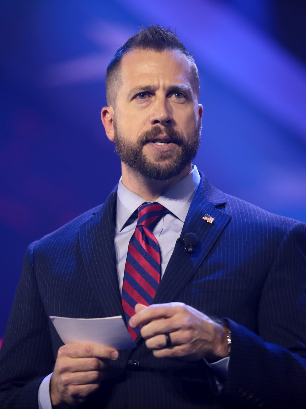 Kyle Seraphin, a bearded man in a navy pinstripe suit and striped tie, speaking on a stage and holding notes.