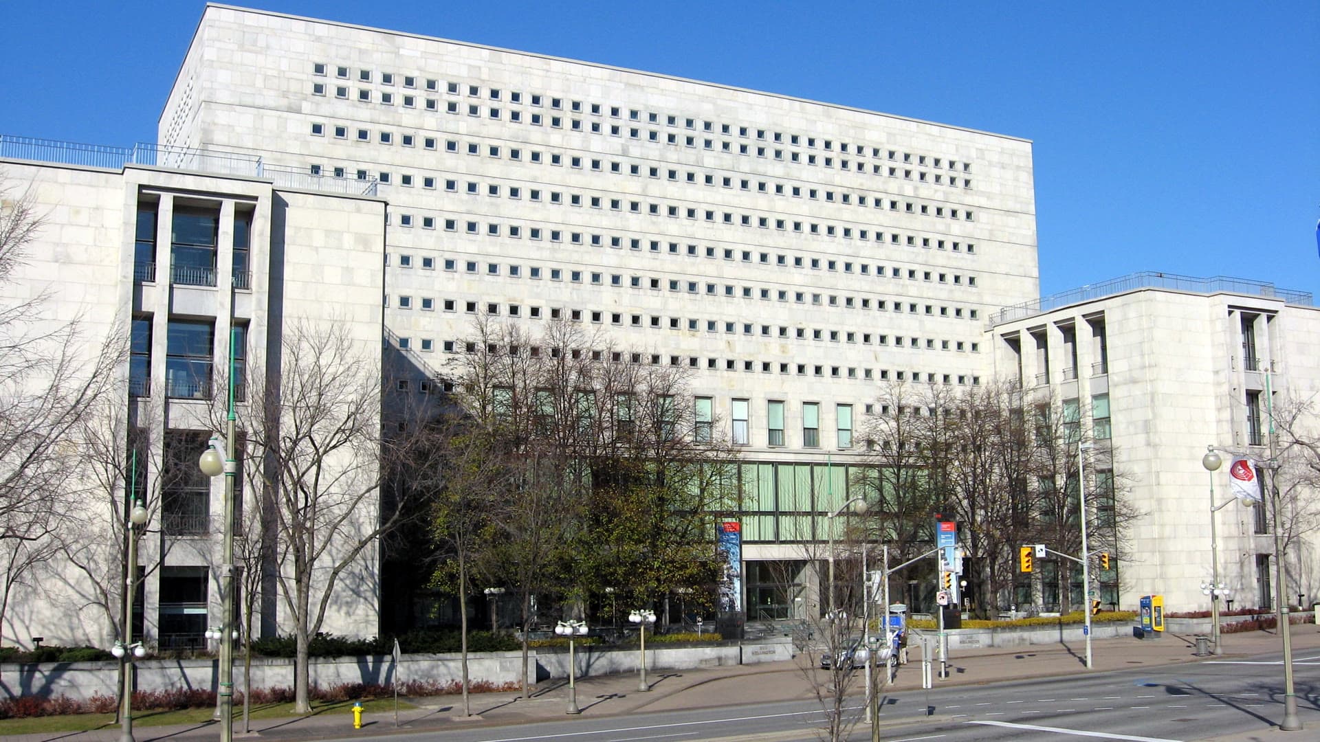 The Wellington Street headquarters of Library and Archives Canada in Ottawa, a pale stone building with rows of small square windows, seen from across the street under a clear blue sky.