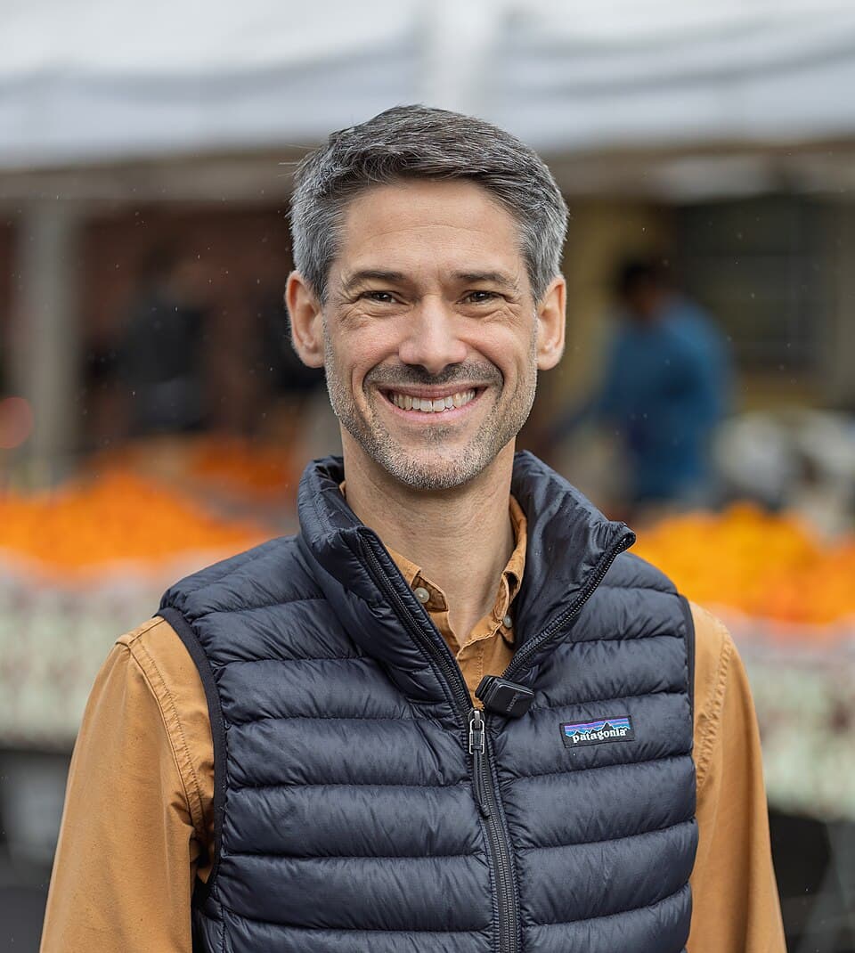 Portrait of Matt Mahan, smiling, wearing a dark puffer vest over a tan shirt at an outdoor farmers market.