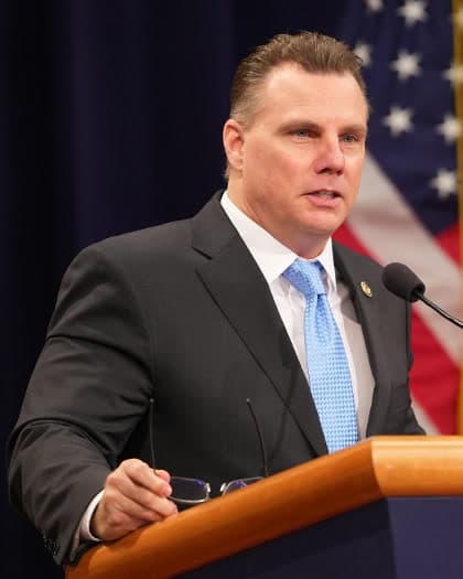 Michael E. Gates, a man with short brown hair in a dark suit, white shirt and light blue tie, speaking at a podium in front of a U.S. flag.