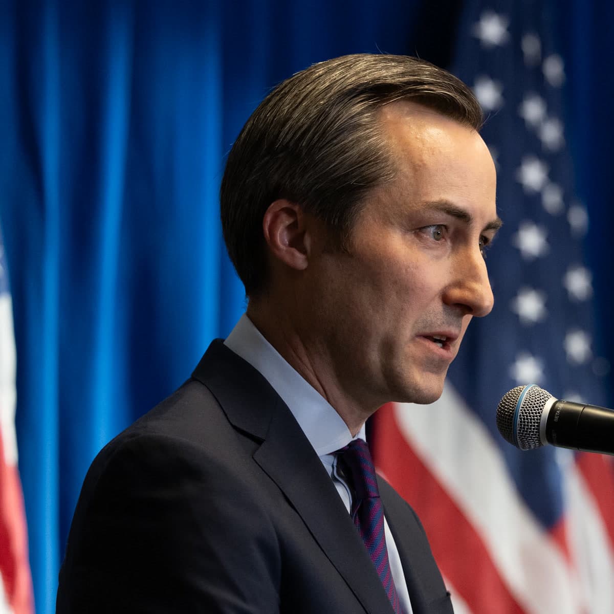 Matthew Miller in a dark suit and purple tie speaking into a microphone in front of American flags and a blue curtain.