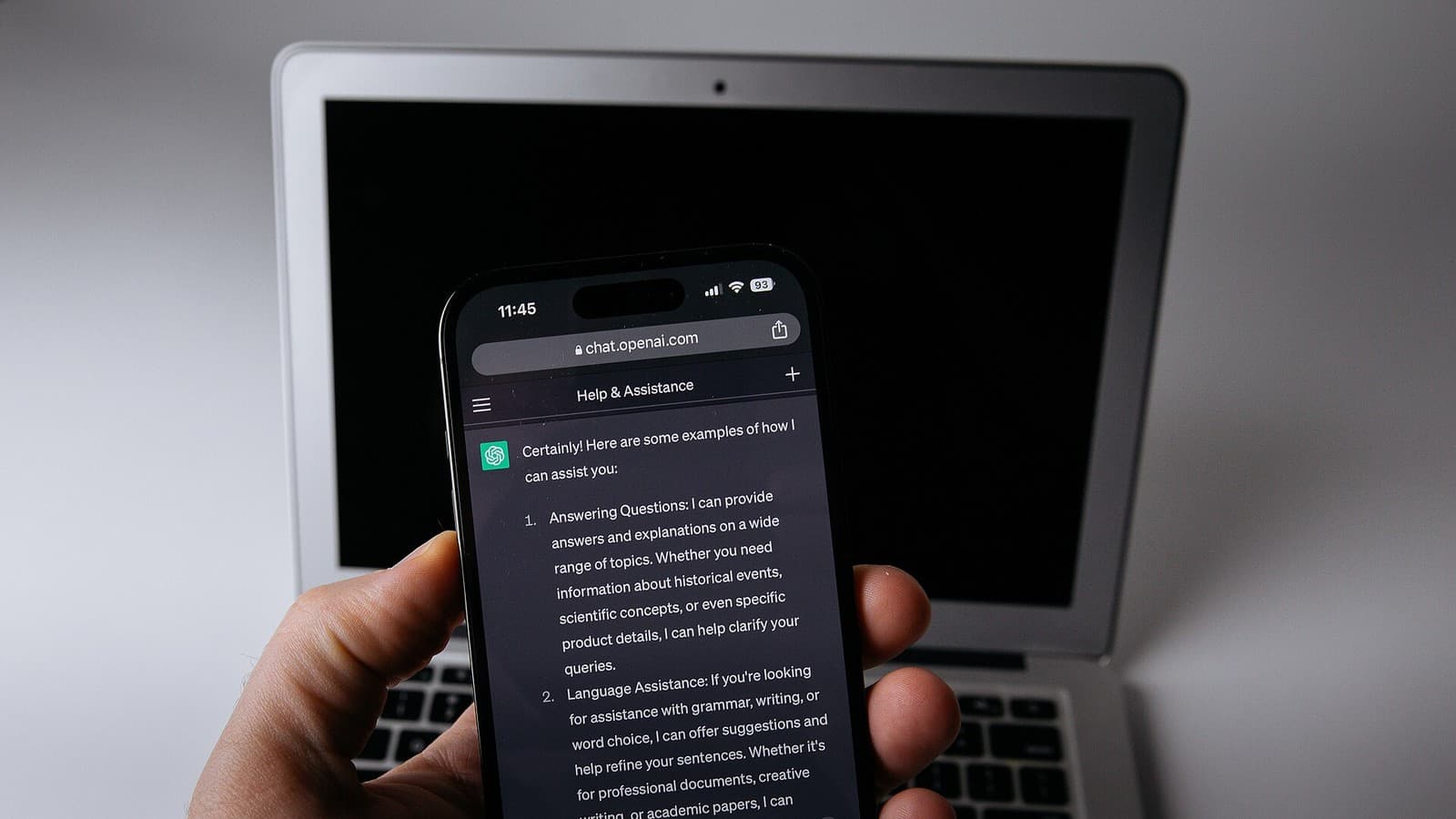 A hand holds a smartphone displaying ChatGPT in front of a laptop.