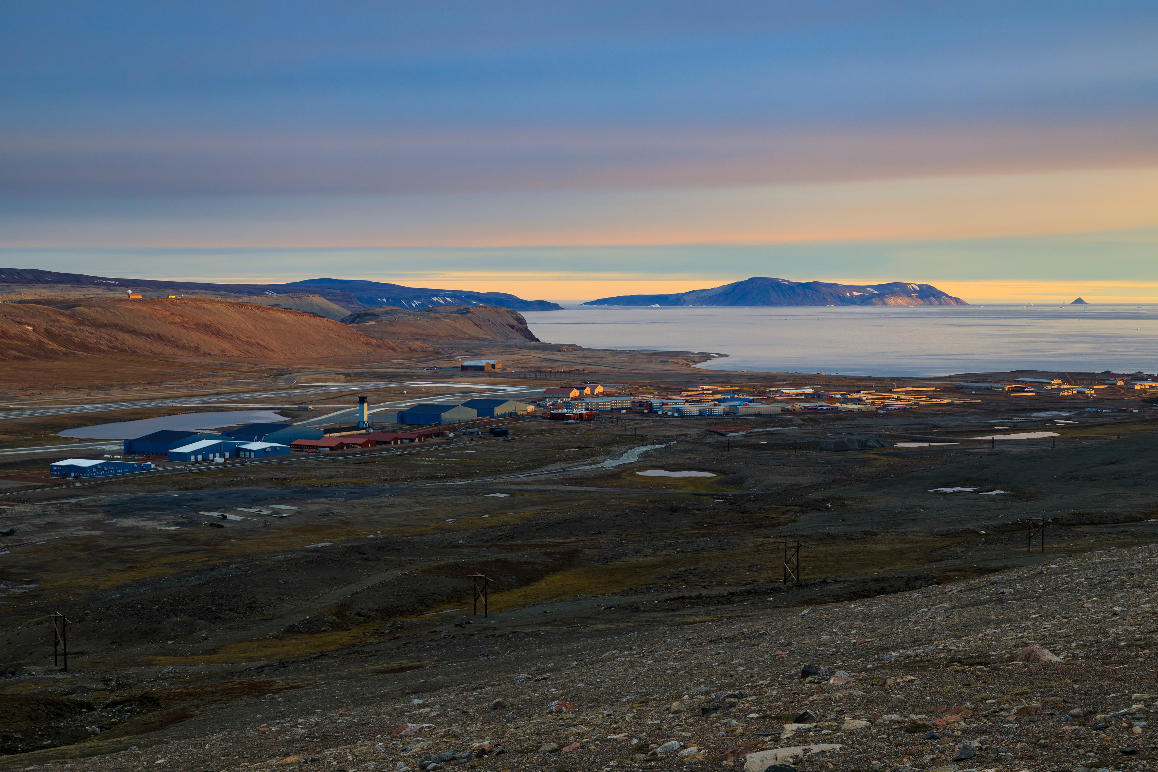 Buildings at Pituffik Space Base beside a Greenland bay and mountains under a pastel sky.