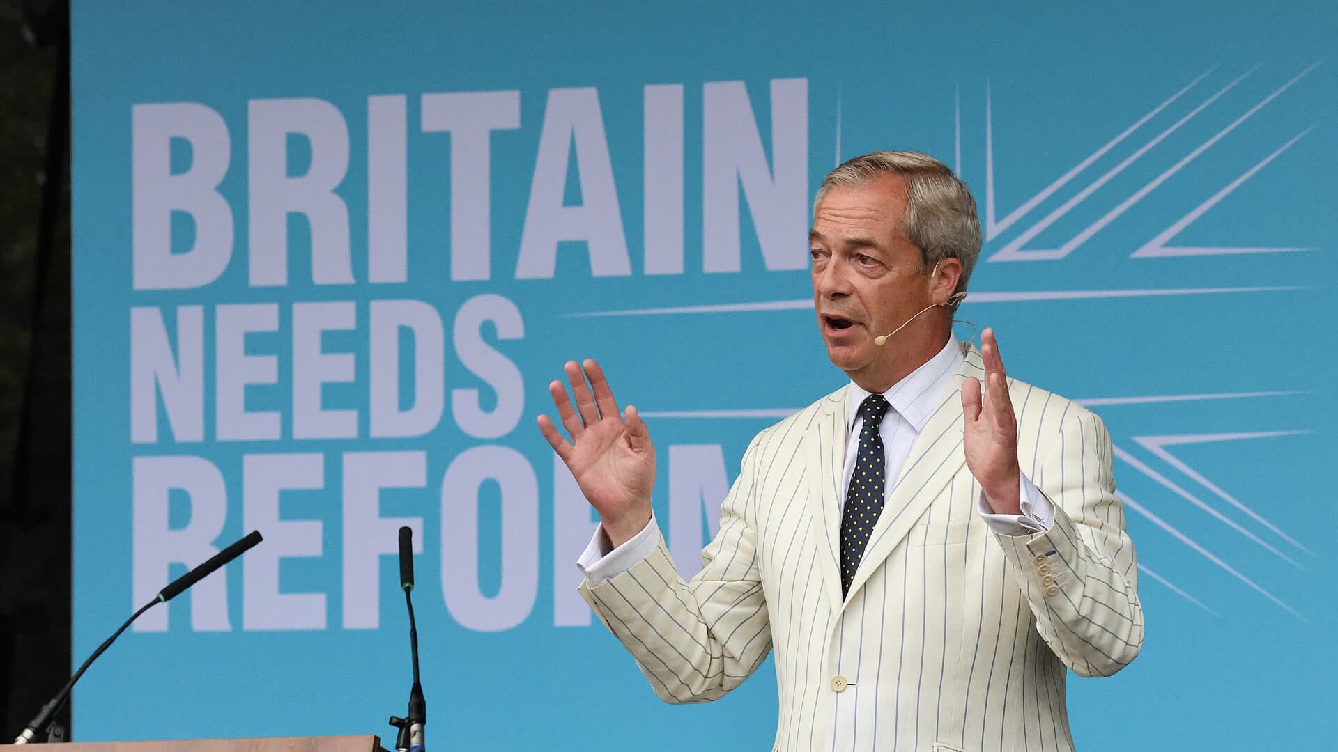 Nigel Farage speaking on stage in front of a turquoise backdrop reading Britain Needs Reform