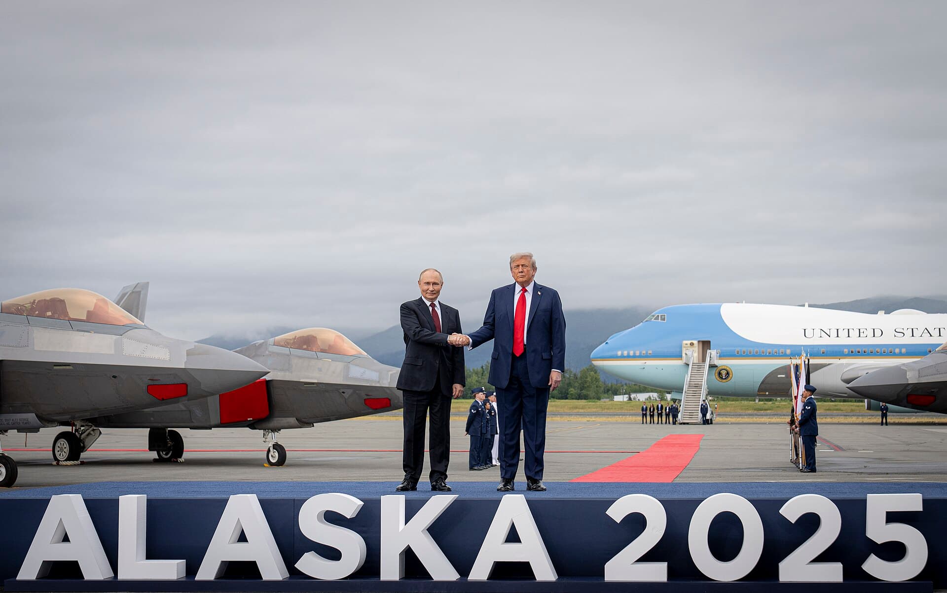 Vladimir Putin and Donald Trump shake hands on a stage marked Alaska 2025, with fighter jets and Air Force One behind them.
