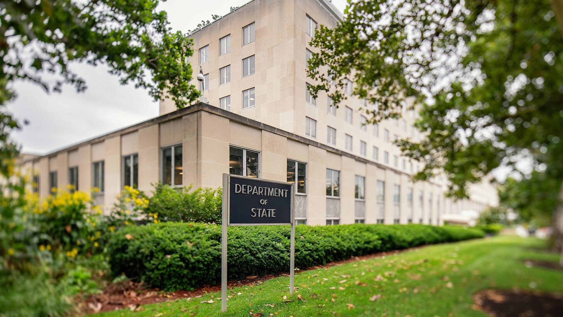 A "Department of State" sign on a lawn in front of the State Department's limestone Harry S. Truman Building in Washington, framed by trees and hedges.