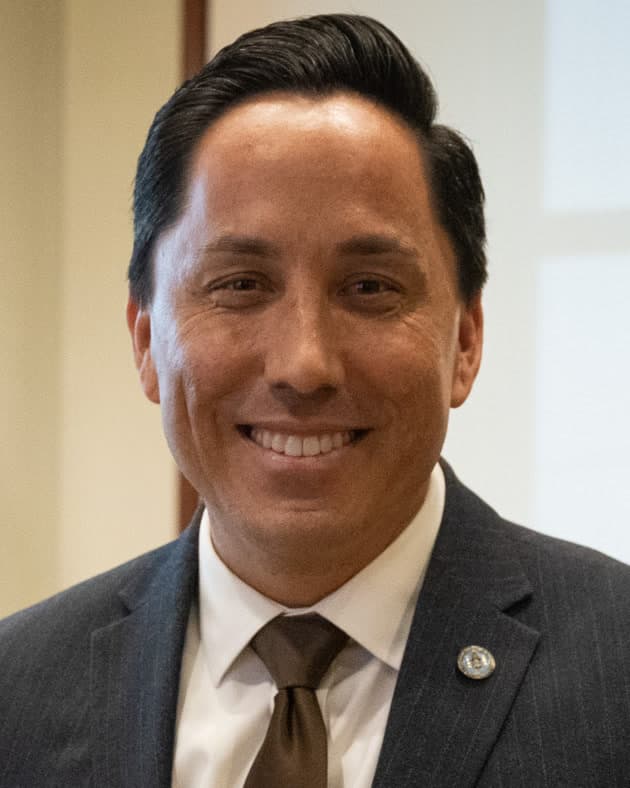 Portrait of Todd Gloria, a man with dark hair, smiling, in a dark pinstriped suit, white shirt and brown tie.