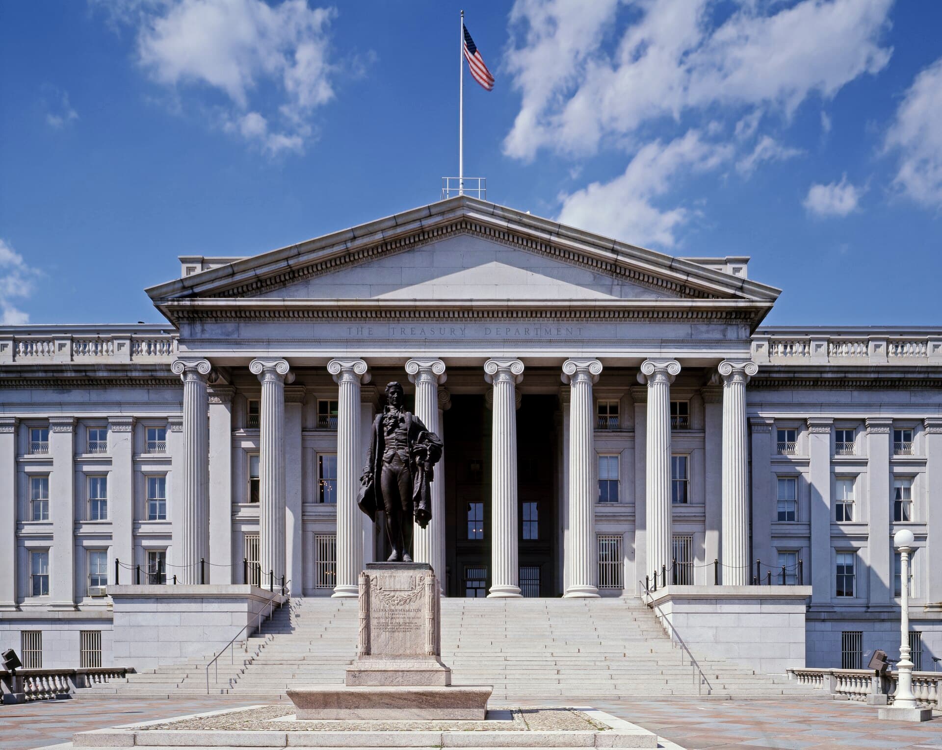 The columned front portico of the U.S. Treasury Building in Washington, inscribed "The Treasury Department," with a bronze statue on a pedestal before broad steps and an American flag on the roof under a blue sky.