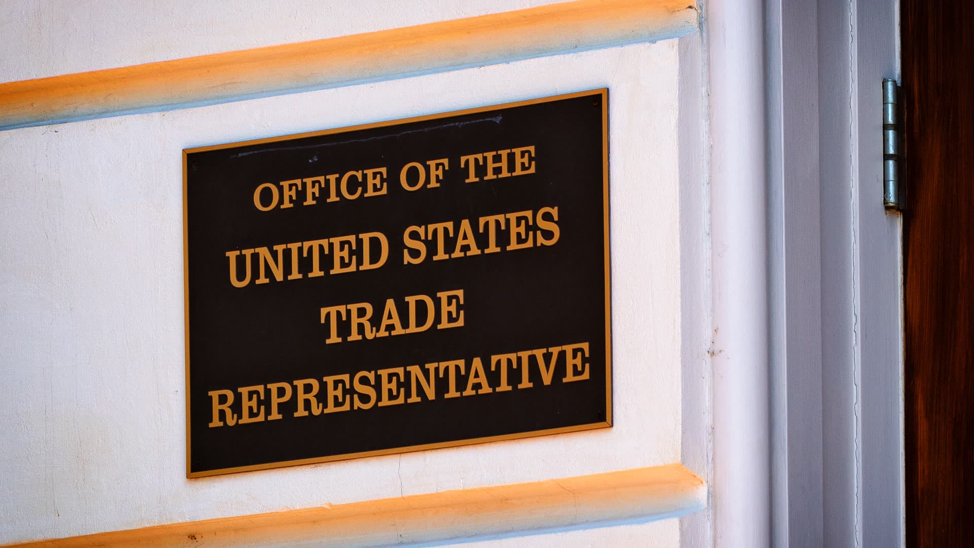 A bronze plaque reading Office of the United States Trade Representative, mounted on a white wall beside a doorway.