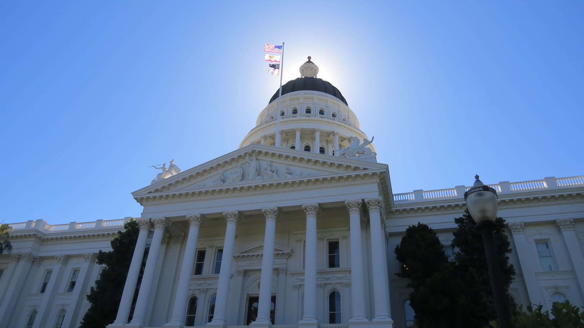 The California State Capitol in Sacramento seen from below, with the dome and flags against a clear blue sky.
