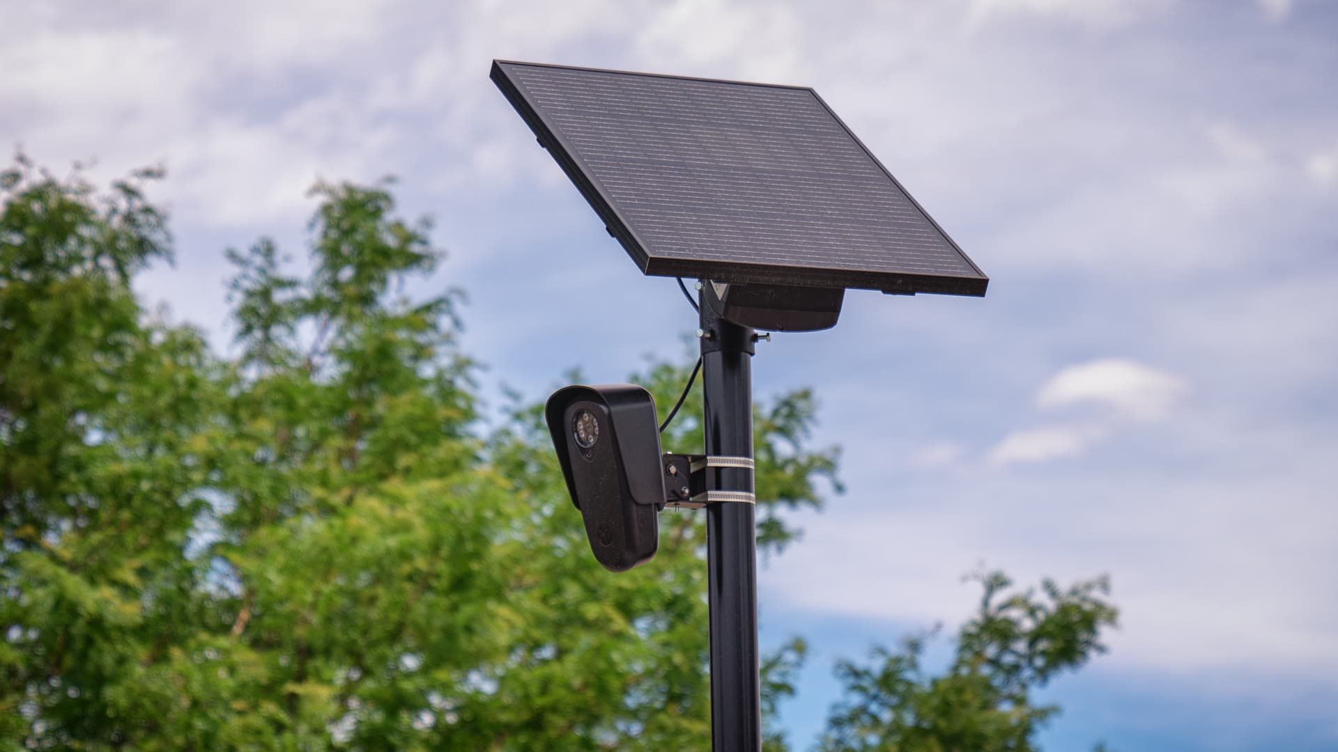 A black Flock Safety license plate reader camera mounted on a pole beneath a solar panel, with trees and a cloudy sky behind it.