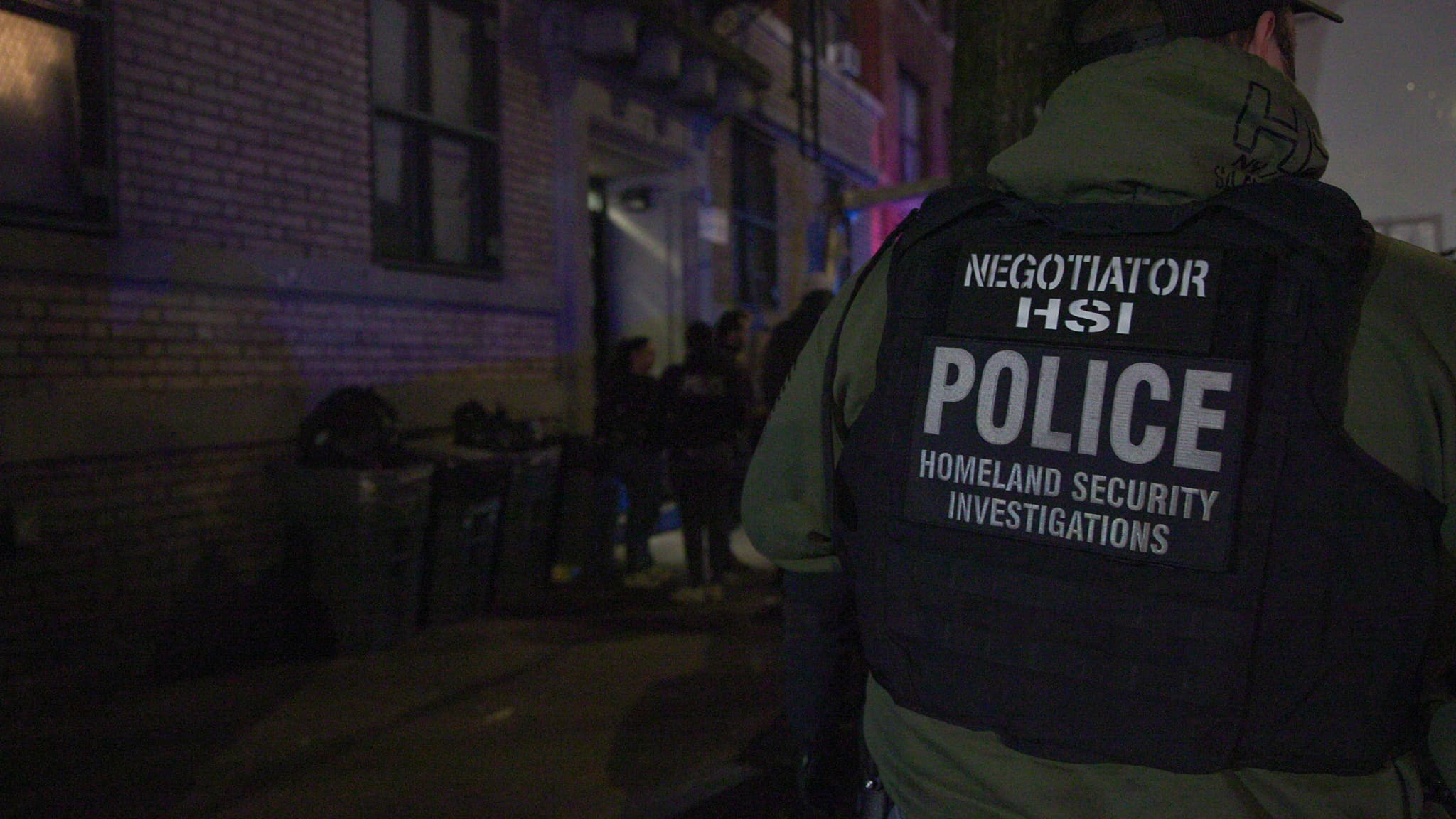 A Homeland Security Investigations agent, seen from behind, outside a Brooklyn apartment building during an arrest-warrant operation at night.