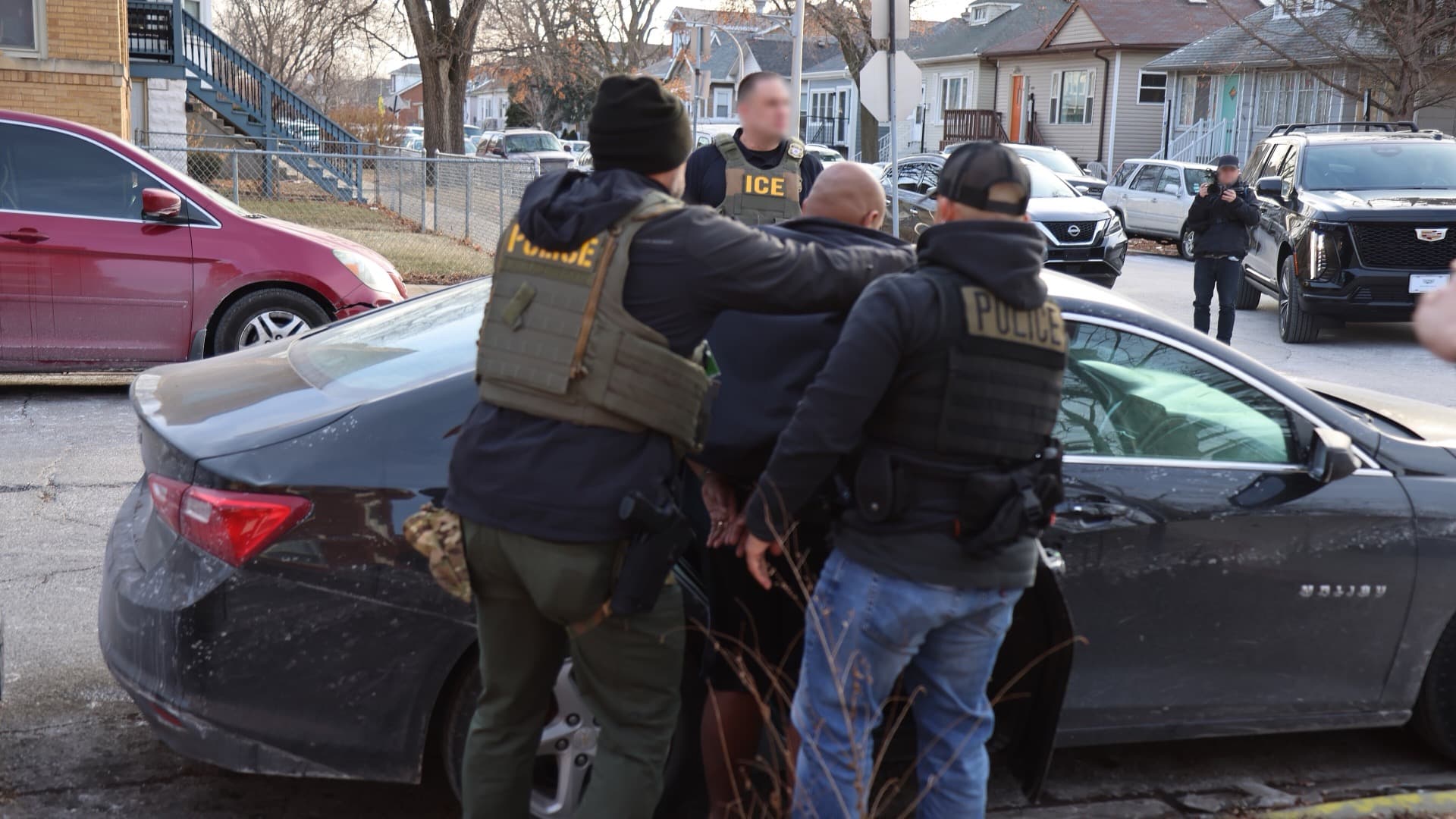Officers in vests marked ICE and POLICE handcuff a man beside a dark sedan on a residential street in Chicago.