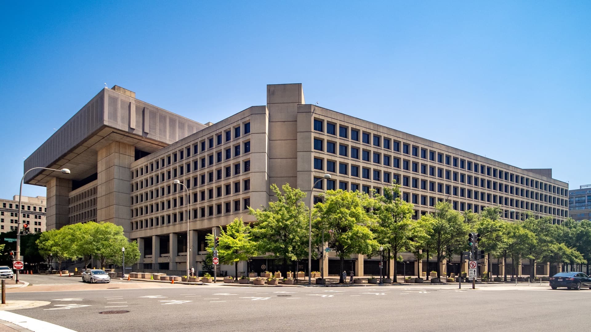 A large beige concrete office building seen from a street corner on a clear day, with green trees along its base.