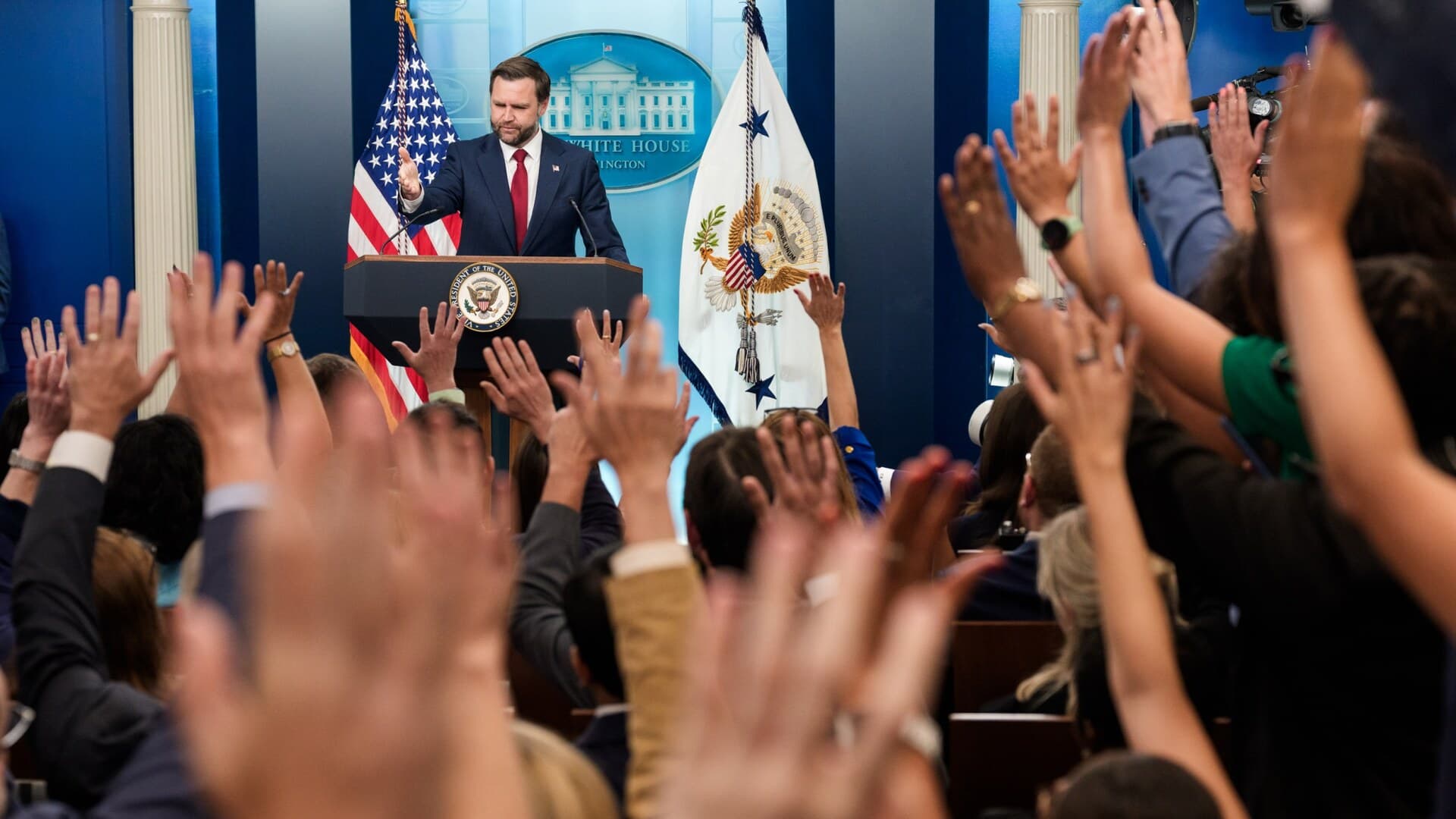 Vice President JD Vance stands at the White House press briefing podium as reporters raise their hands.