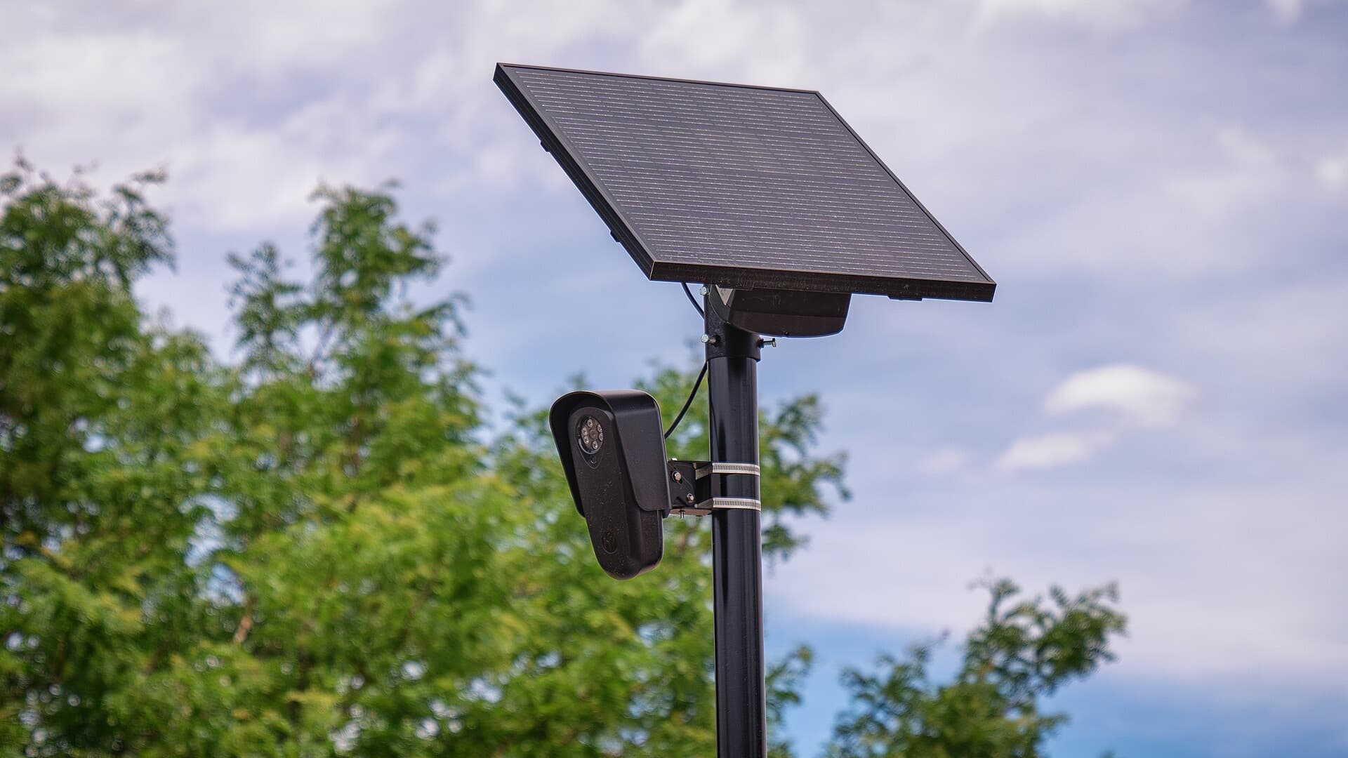 A black Flock Safety license-plate reader camera mounted on a pole under a solar panel, with trees and a cloudy sky behind it.