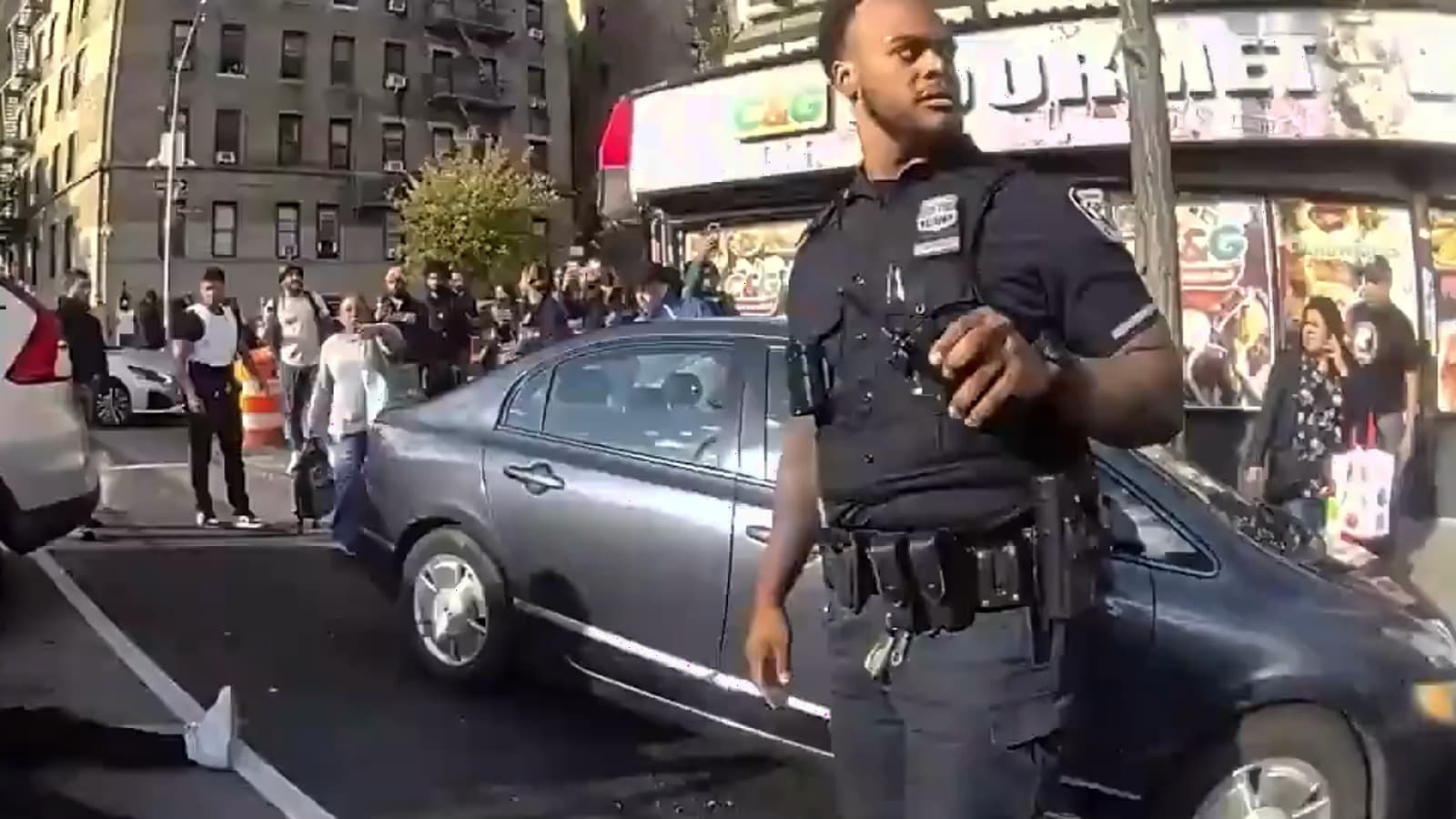 An NYPD officer stands near vehicles and bystanders at the Marble Hill shooting scene.