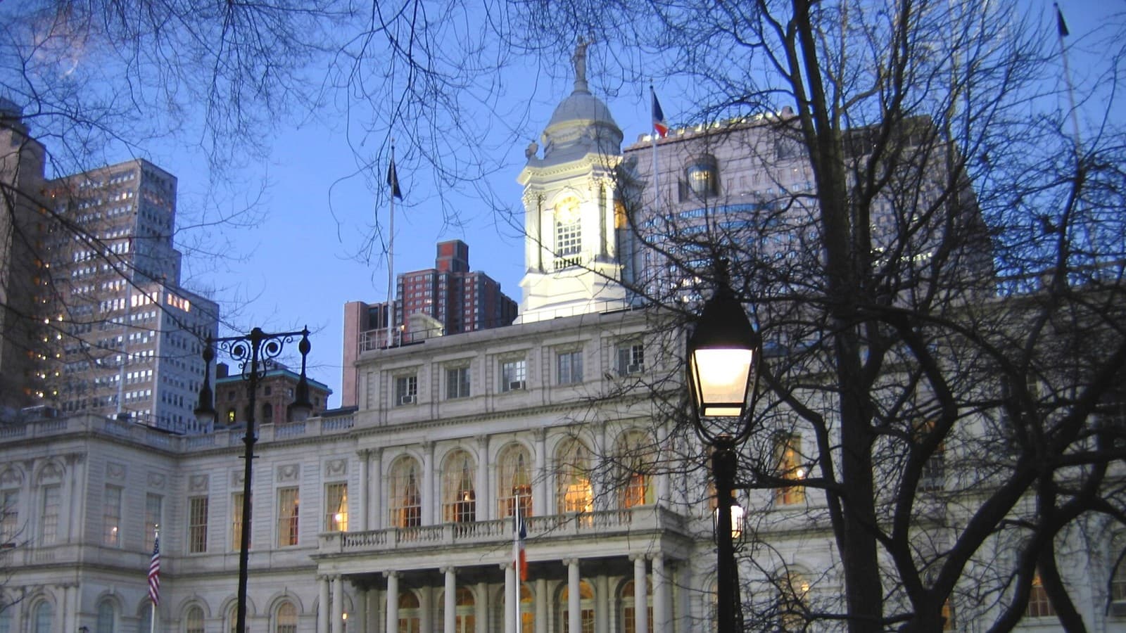 New York City Hall illuminated at dusk behind trees and a streetlamp.