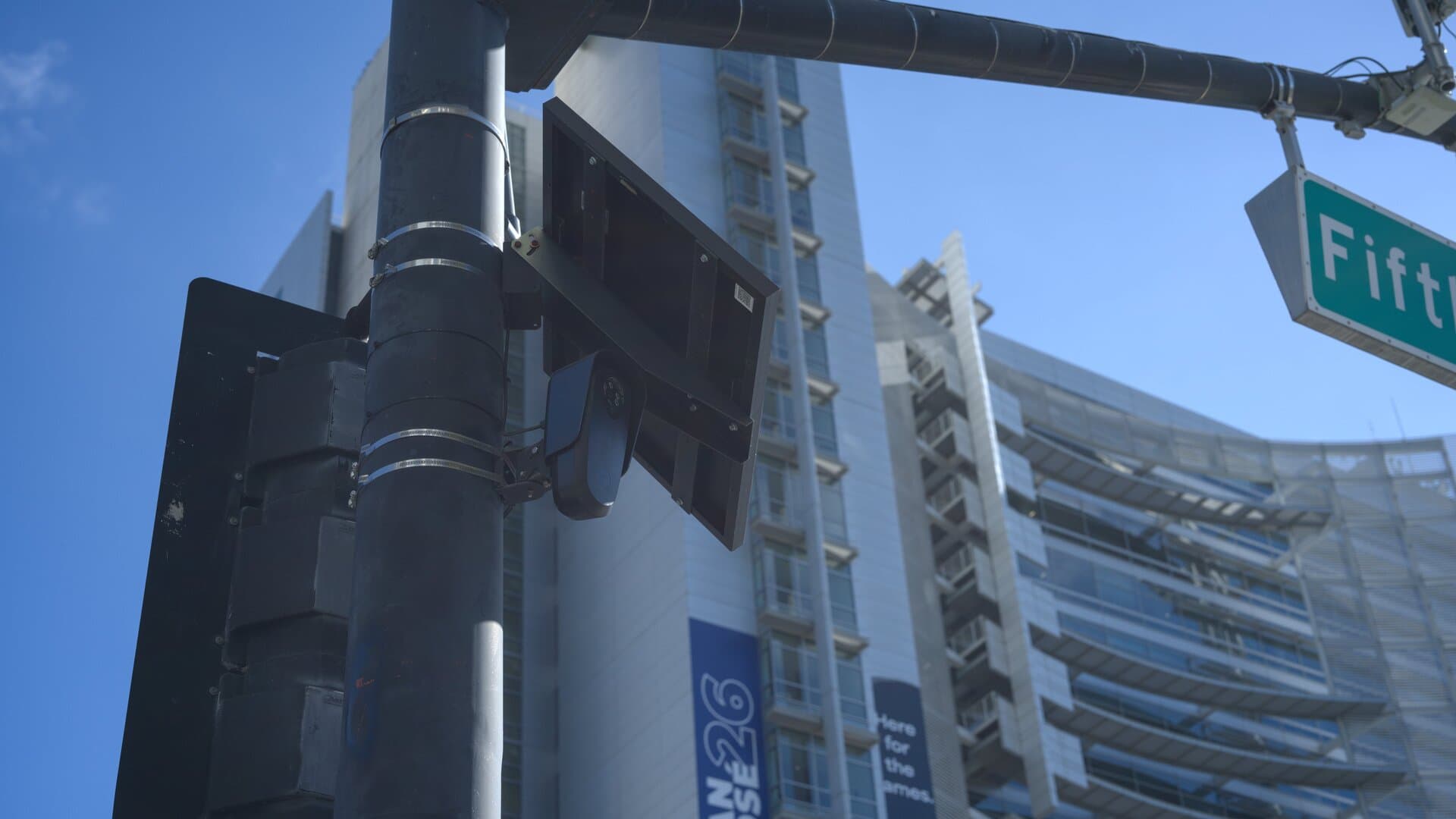 A small black Flock Safety license plate reader camera mounted under a solar panel on a traffic signal pole at East Santa Clara and North Fifth streets in downtown San Jose, with San Jose City Hall and a Fifth St street sign behind it.